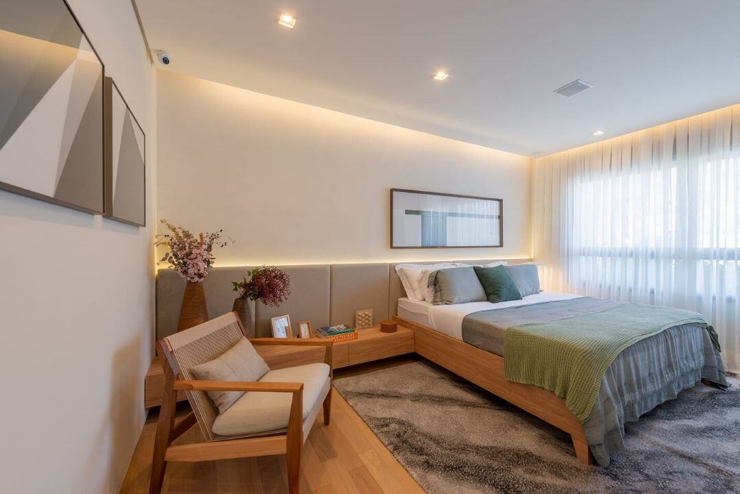 Foto da decoração de quarto de casal com paredes bege e móveis em madeira e janela com cortina branca.