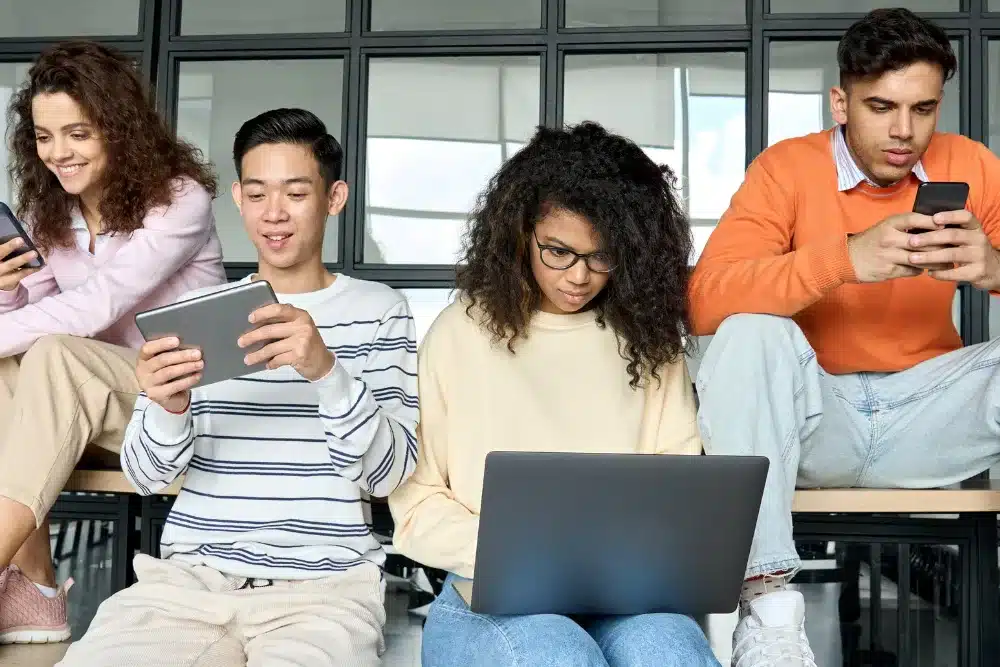 Esta imagem mostra quatro jovens usando dispositivos digitais em um ambiente que se assemelha a um escritório ou sala de aula moderna