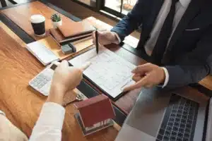Foto de duas pessoas negociando um imóvel, com um notbook e alguns papéis em cima da mesa de madeira.