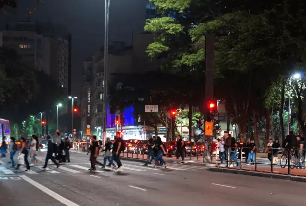 Uma via urbana movimentada, mostrada à noite com tráfego de pedestres e carros, e edifícios ao redor.